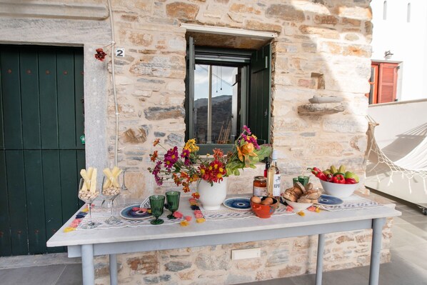 A table set with flowers, fruits, and snacks outside a stone building with a green door and a window.
