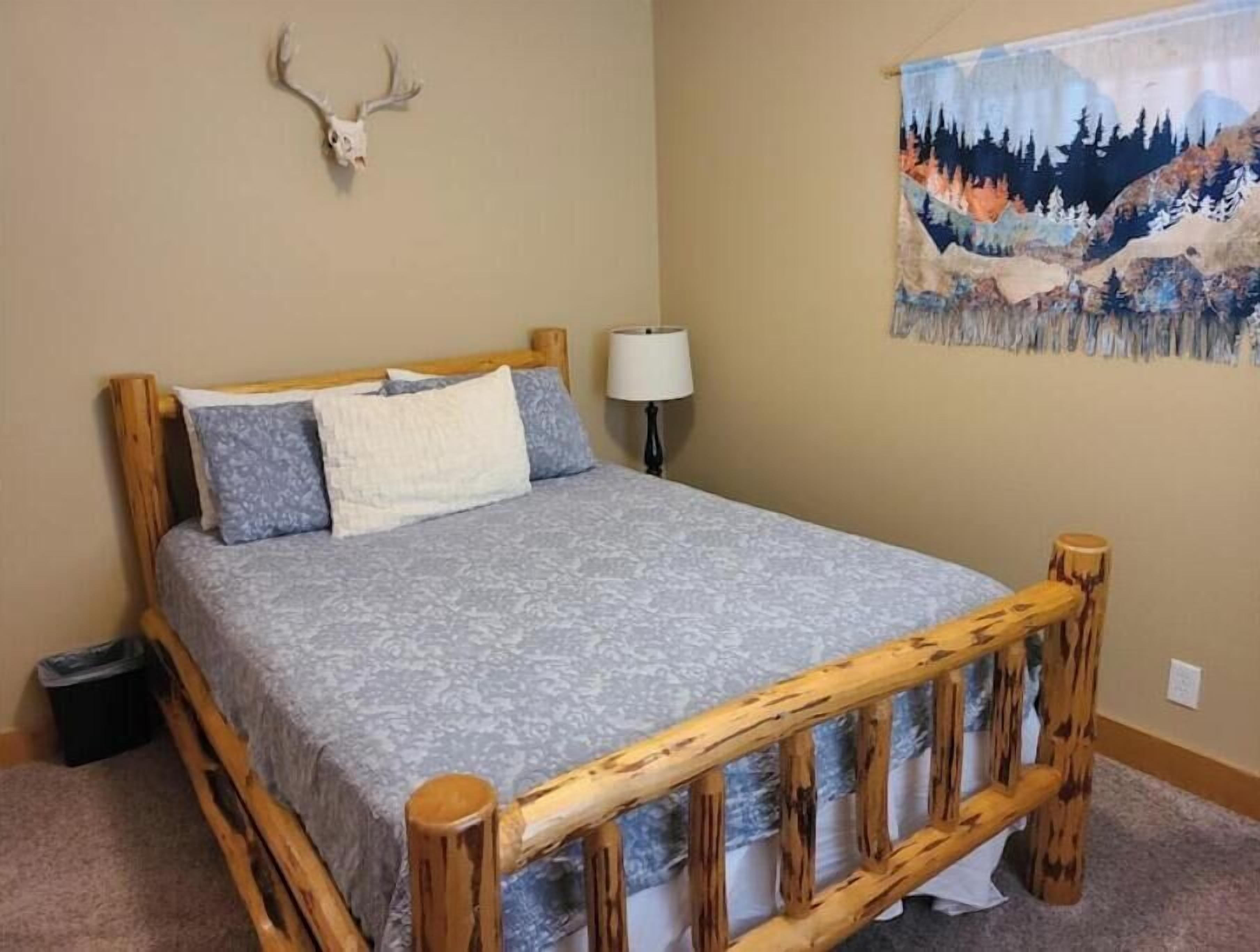 A bedroom with a wooden bed frame, a patterned bedspread, a wall-mounted lamp, and a painting of a forest scene.