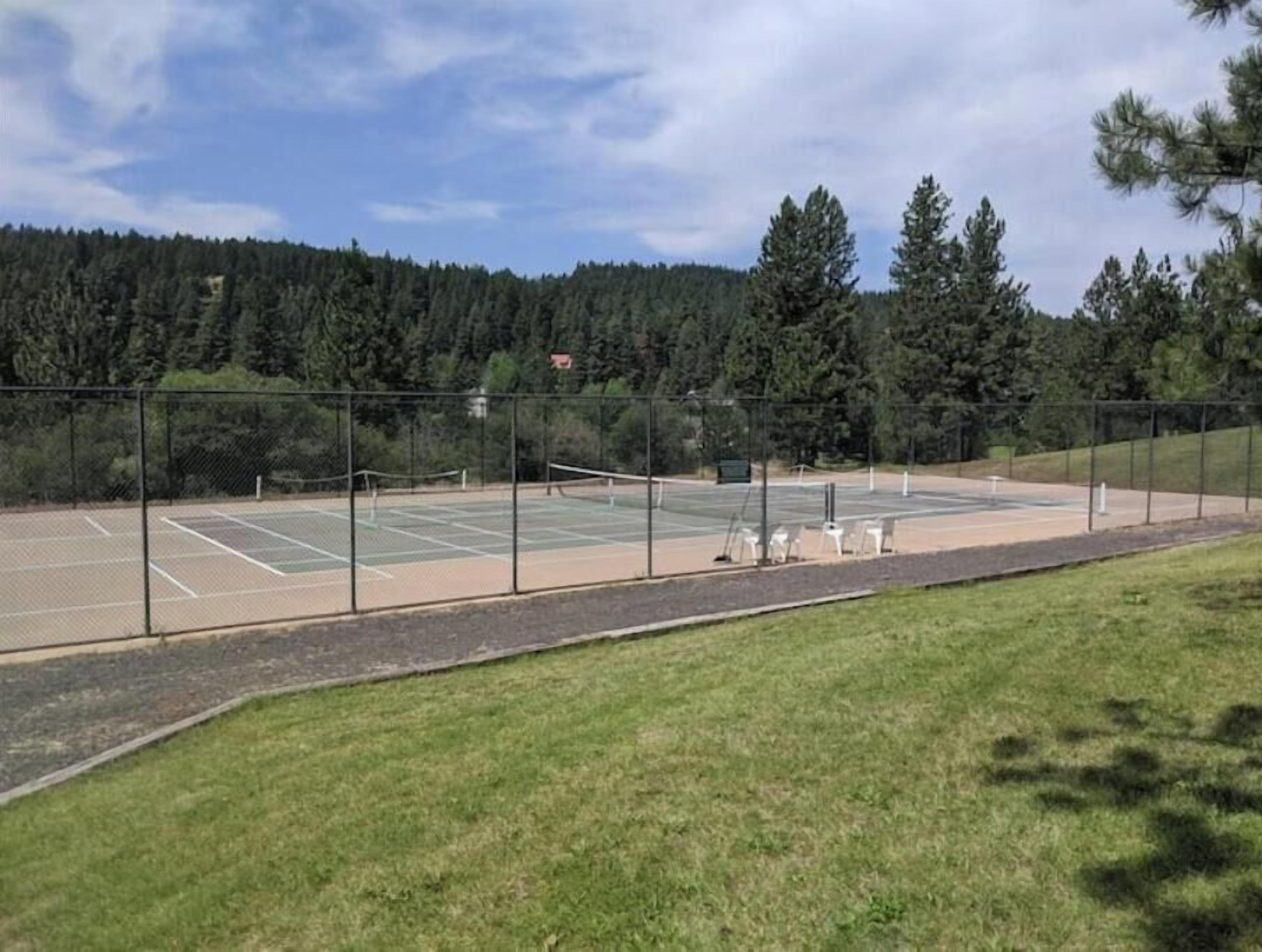 An outdoor tennis court with a fence, surrounded by grass and trees.