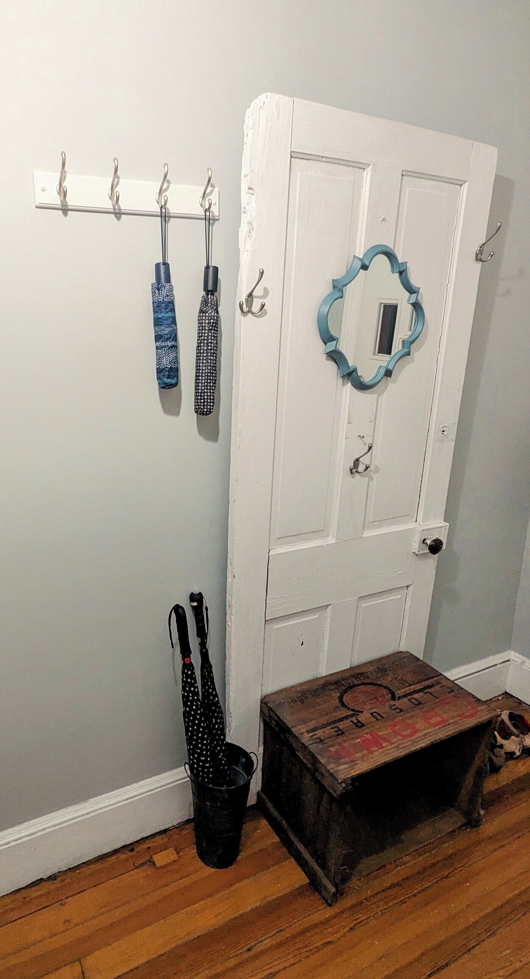 Una puerta blanca con un espejo azul, un perchero con paraguas y un caja de madera en el suelo.