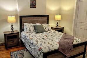 Un dormitorio con estructura de cama de madera, ropa de cama floral, dos lámparas de noche, una mesita de noche y una puerta.