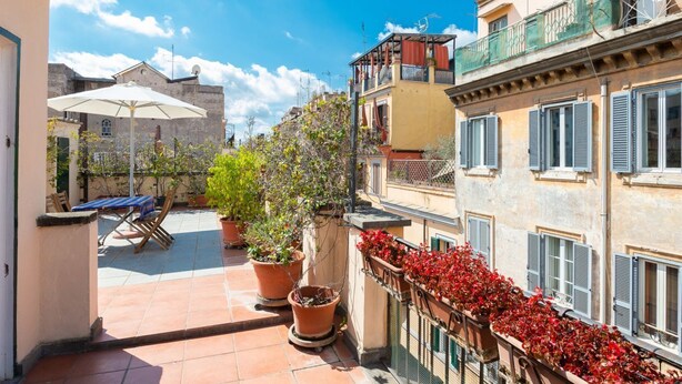 A courtyard with a table and chairs, potted plants, and a balcony with red flowers. - Fori Imperiali Amazing Terrace (Roma)