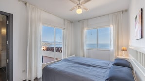 Une chambre à coucher comprenant un lit, deux tables de chevet, un ventilateur de plafond et de grandes fenêtres donnant sur la mer.