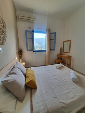 A bedroom with a bed, a window with blue shutters, a wooden nightstand, a mirror, and a wall-mounted air conditioner.