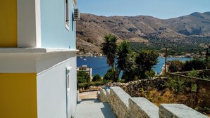 A coastal view with a stone wall, a building with a blue facade, and a mountainous landscape in the background. - Apartment 'Irene Family' with Sea View, Wi-Fi and Air Conditioning (Symi)