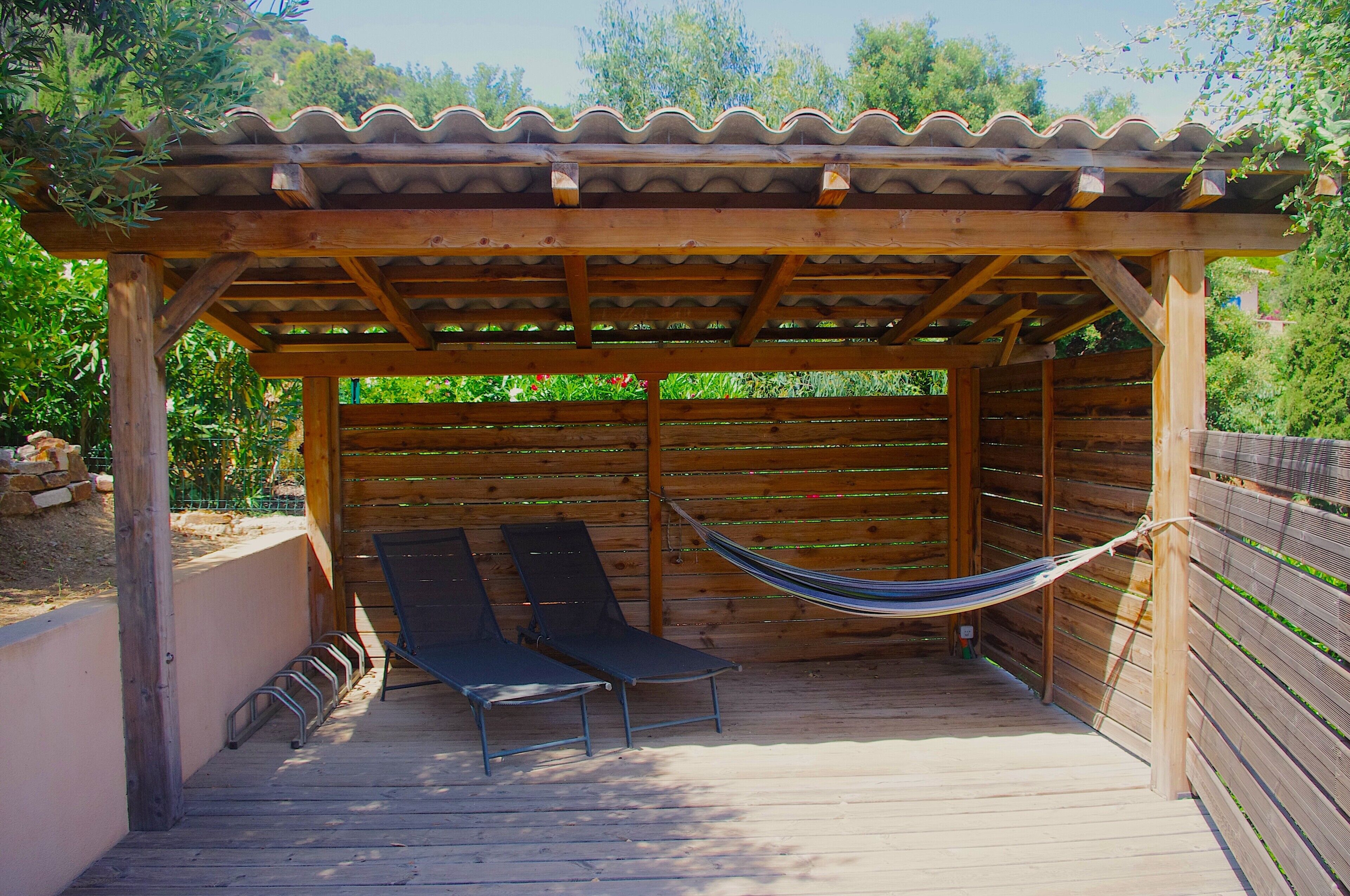 A wooden pergola with a tiled roof, two lounge chairs, and a hammock.