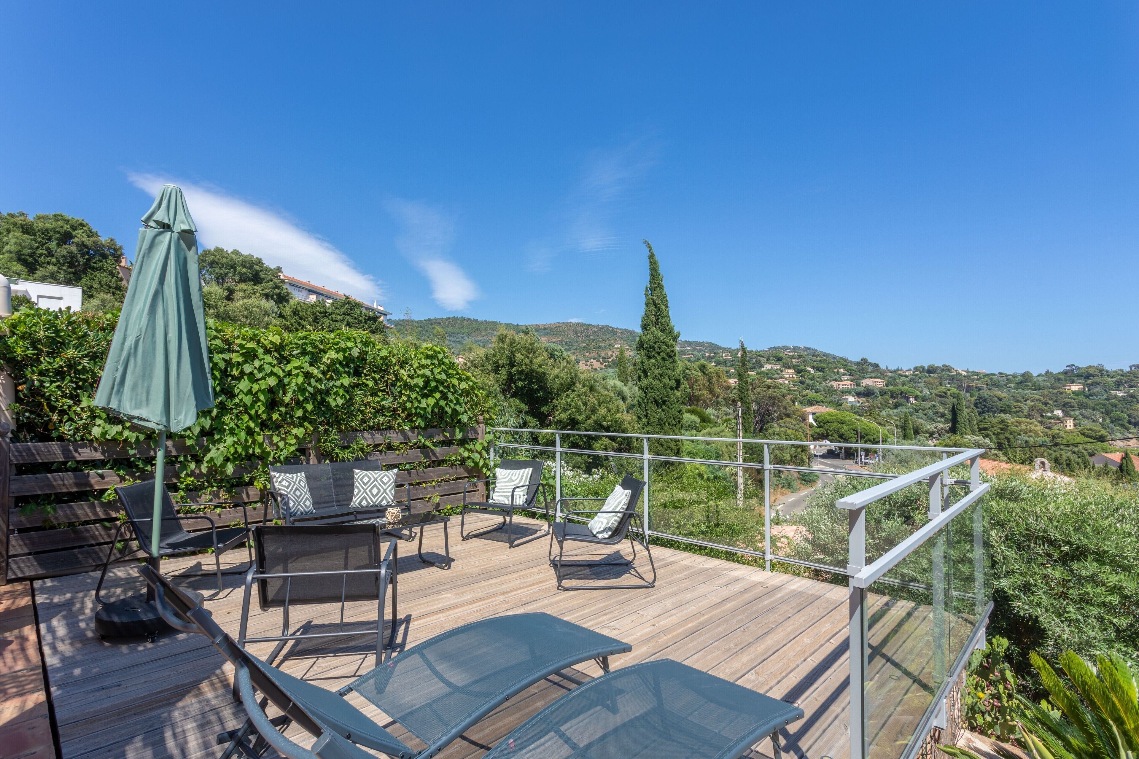 Um deck de madeira com móveis de exterior, um guarda-sol verde e vista para uma paisagem montanhosa.