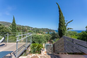 A wooden deck with a metal railing, overlooking a coastal landscape with trees and a view of the sea.