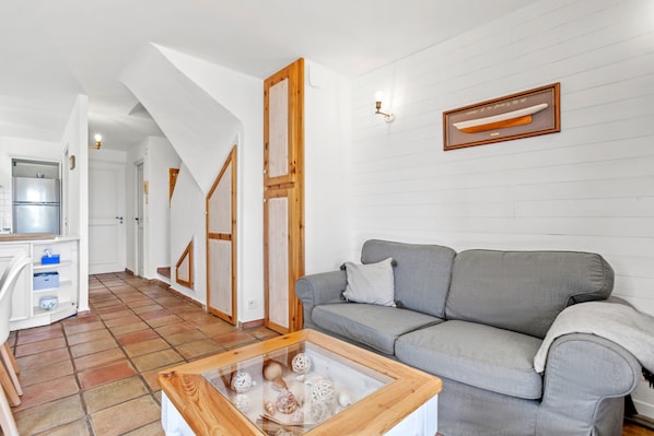 Un salón con un sofá gris, una mesa de centro de madera con adornos, una zona de cocina con refrigerador y un cuadro en la pared.