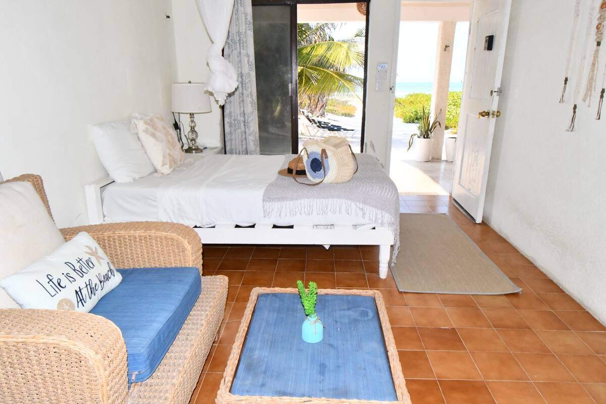 Une chambre avec un lit, deux fauteuils en osier, une petite table avec une plante, et une vue sur la plage par une porte ouverte.
