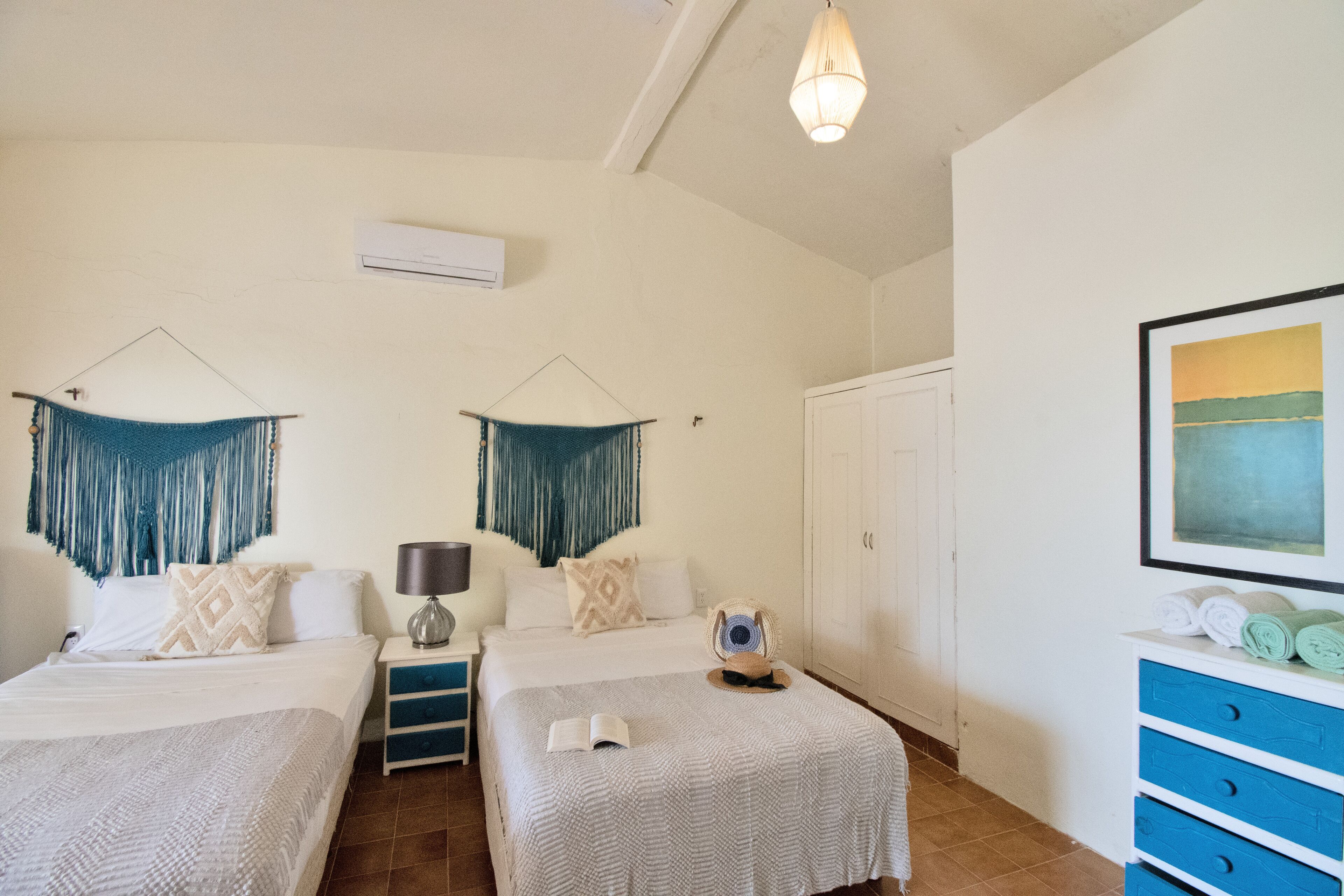 A hotel room with two beds, a wardrobe, a desk with a lamp, and a painting on the wall.
