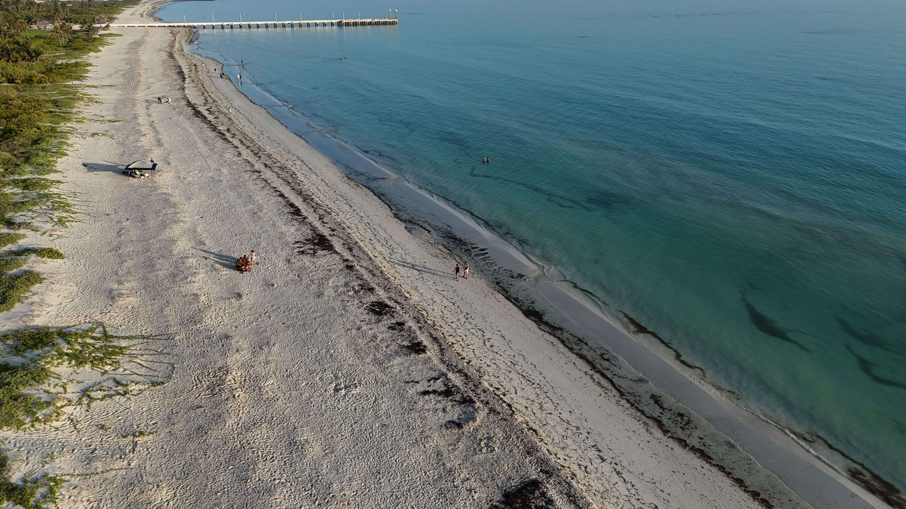 A beach with clear turquoise water, a sandy shore, and a pier in the distance.