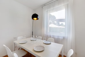 Un comedor con una mesa blanca, sillas, una lámpara colgante negra y una ventana con cortinas translúcidas.