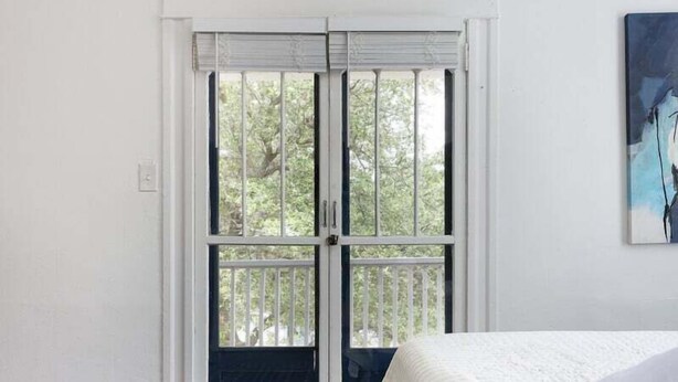 Un dormitorio con cama, alfombra estampada, puerta blanca con vistas y un cuadro en la pared.