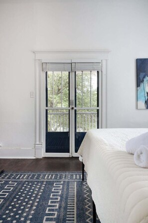 Une chambre à coucher avec un lit, un tapis à motifs, une porte blanche donnant sur un paysage et un tableau accroché au mur.