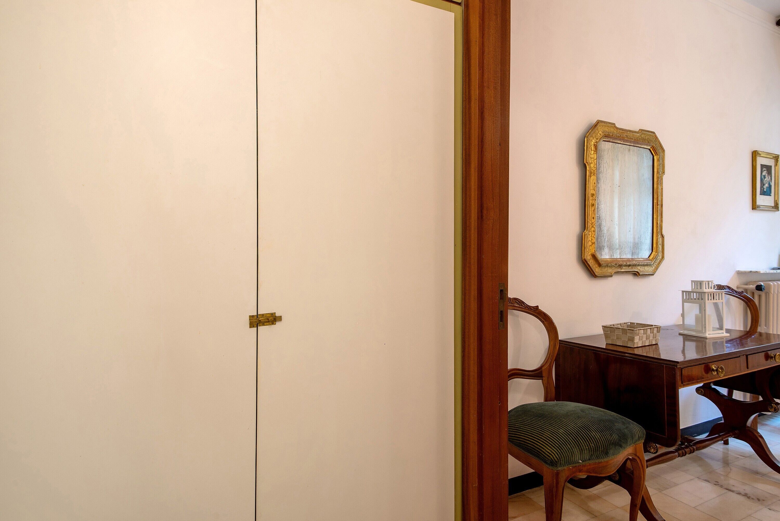 A room with a closed door, a wooden chair, a desk with a mirror, and a framed picture on the wall.