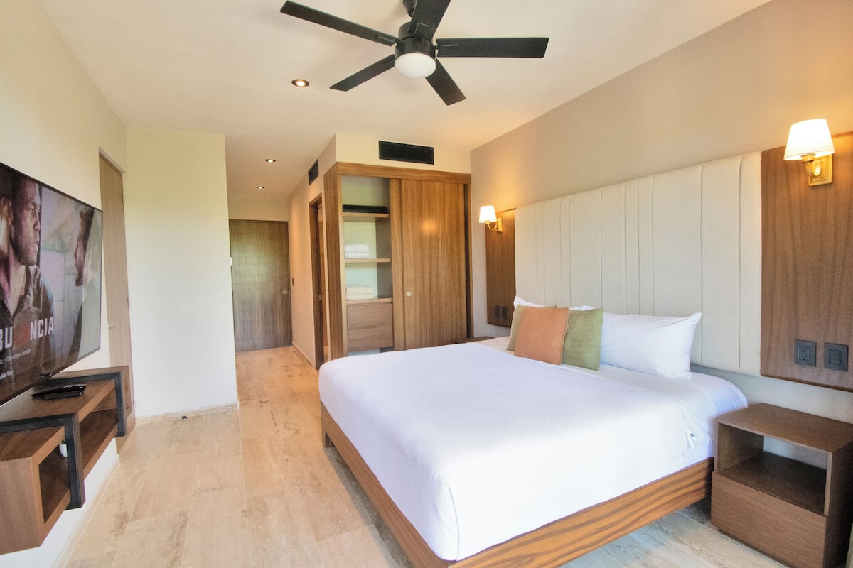 Une chambre d’hôtel avec un grand lit, une tête de lit en bois, un ventilateur de plafond, des lampes de chevet et un téléviseur posé sur un meuble en bois.