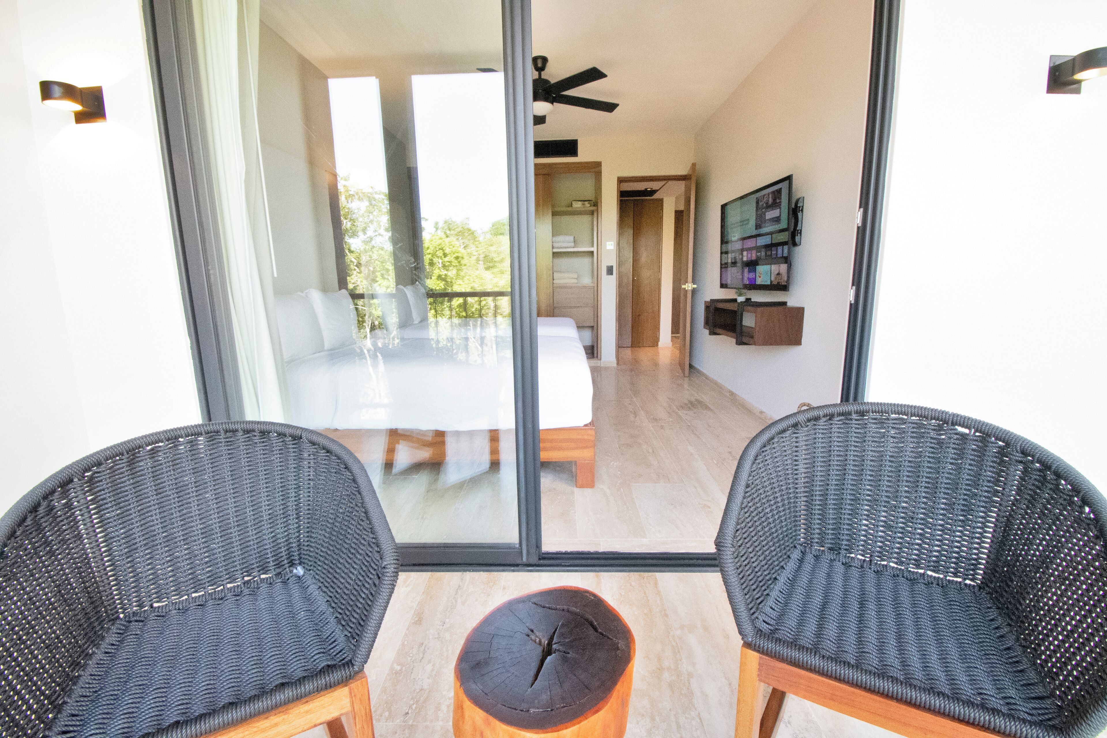 A balcony with two wicker chairs, a small wooden stool, and a glass door leading to a dining area with a table and chairs.
