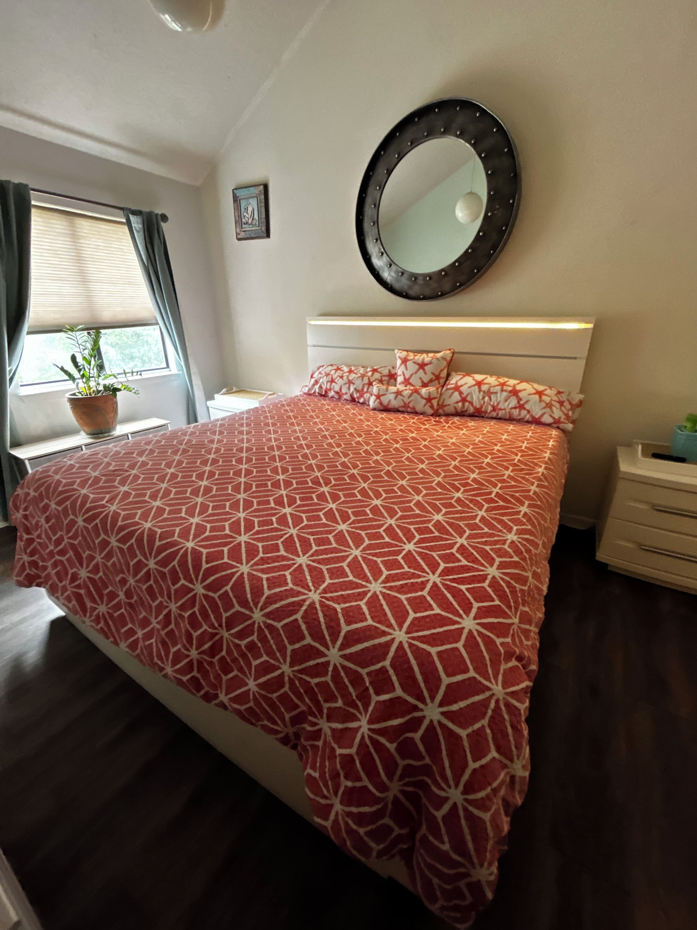 A bedroom with a large bed, a round mirror, a potted plant, and a window with curtains.
