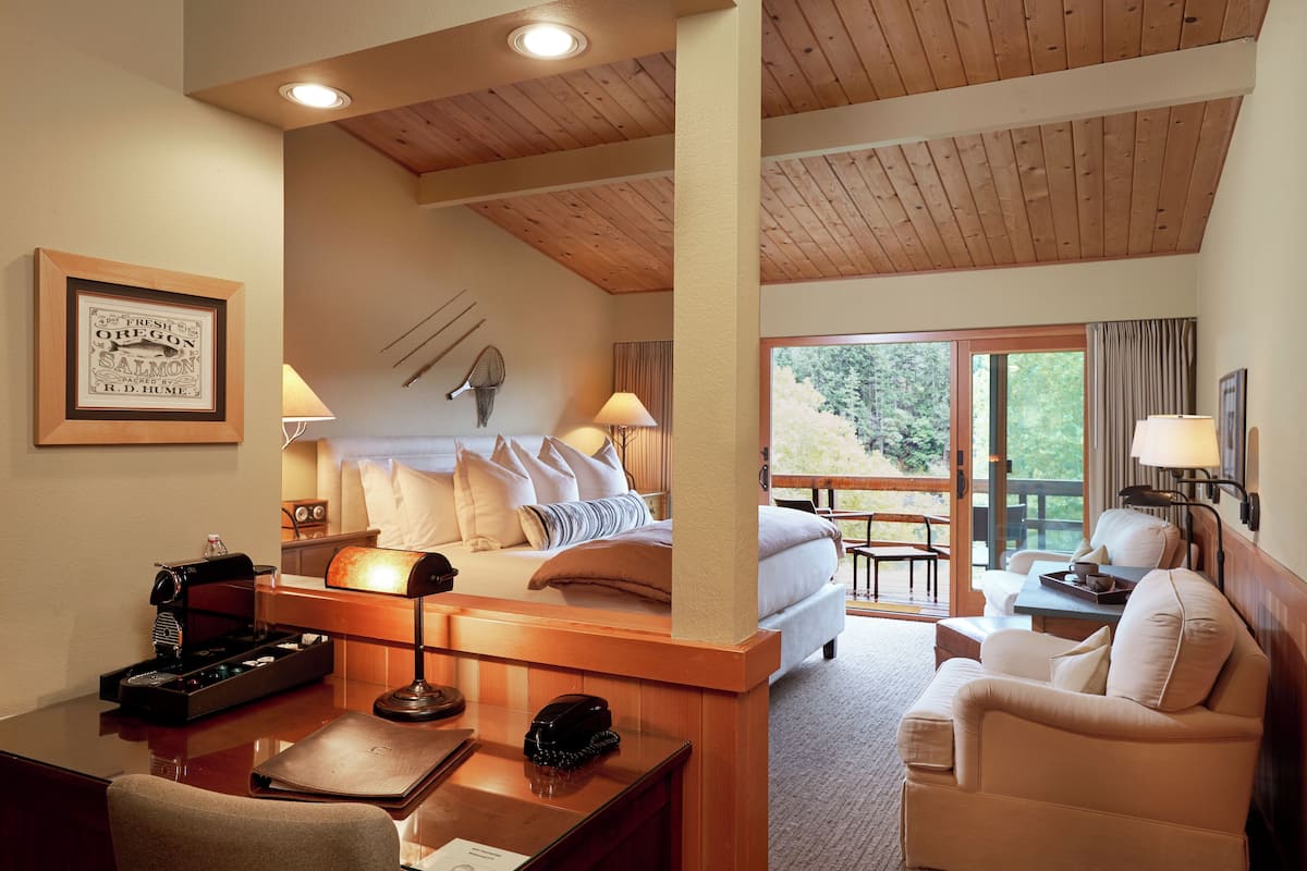 Habitación de hotel con techo de madera, cama con ropa blanca, escritorio con lámpara, una silla y vistas al exterior a través de una puerta de cristal.