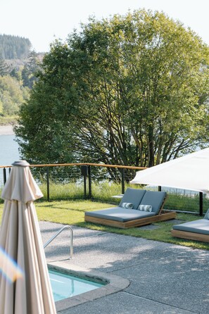 Un espace aménagé au bord de la piscine, avec des chaises longues et un parasol, entouré d’arbres et offrant une vue sur l’eau.