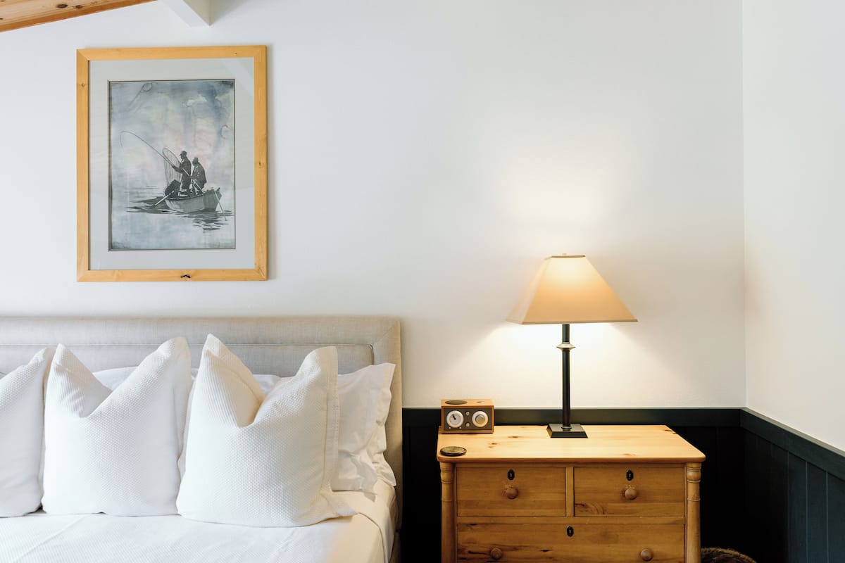 Una cama bien hecha con ropa de cama y almohadas blancas, una obra enmarcada que representa una escena con barcos, una mesita de noche con una lámpara y un radio, y un mueble de noche de madera con cajones.