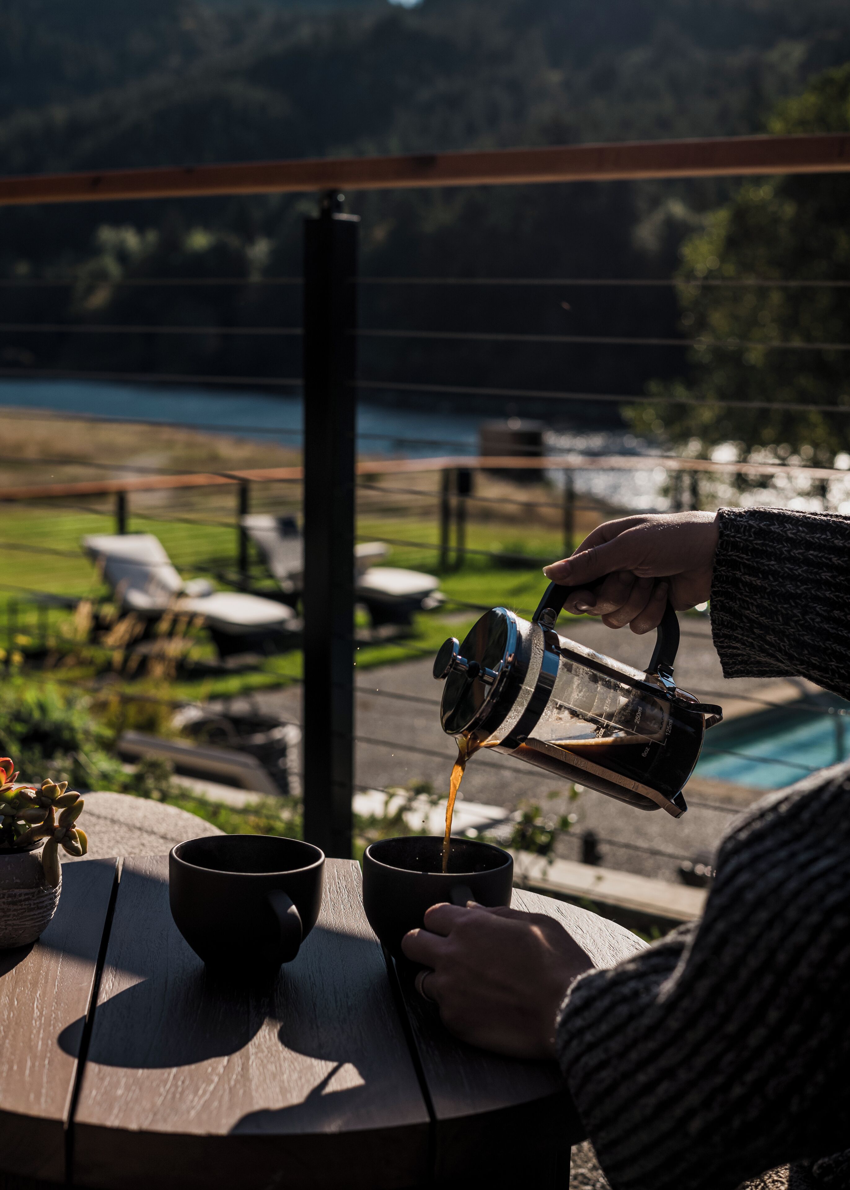 A person pouring coffee into a cup on a wooden table outdoors with a pool and chairs in the background.
