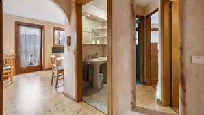 Una habitación con piso de madera, una silla, una puerta que da a un baño con lavamanos e inodoro, y una ventana con cortina.