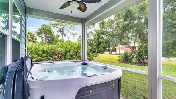 Ein Whirlpool in einem überdachten Außenbereich mit Deckenventilator, umgeben von Grünpflanzen und Blick auf ein Haus.