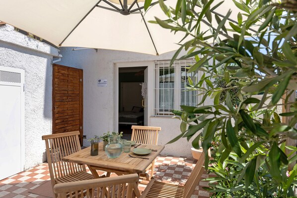 Un patio al aire libre con una mesa y sillas de madera, una pared blanca, una puerta de madera y una ventana con barrotes blancos.