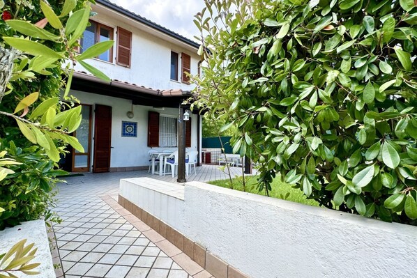 A house with a tiled patio, a dining area with chairs, and a garden with green foliage.