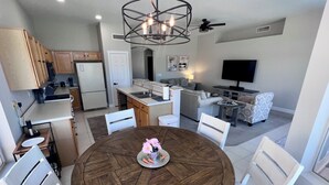 A modern kitchen with wooden cabinets, a dining table with chairs, a chandelier, and a living area with a sofa and TV. - Desert Oasis / Heated Pool / Spring Training / Barrett-Jackson / WM Phoenix Open (Surprise)