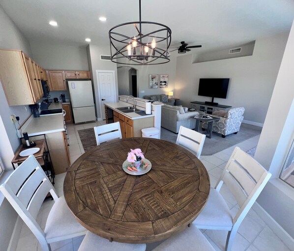 A modern kitchen with wooden cabinets, a dining table with chairs, a chandelier, and a living area with a sofa and TV.