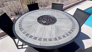 A round outdoor table with a central fire feature, surrounded by wicker chairs, overlooking a swimming pool and a landscaped yard. - Desert Oasis / Heated Pool / Spring Training / Barrett-Jackson / WM Phoenix Open (Surprise)