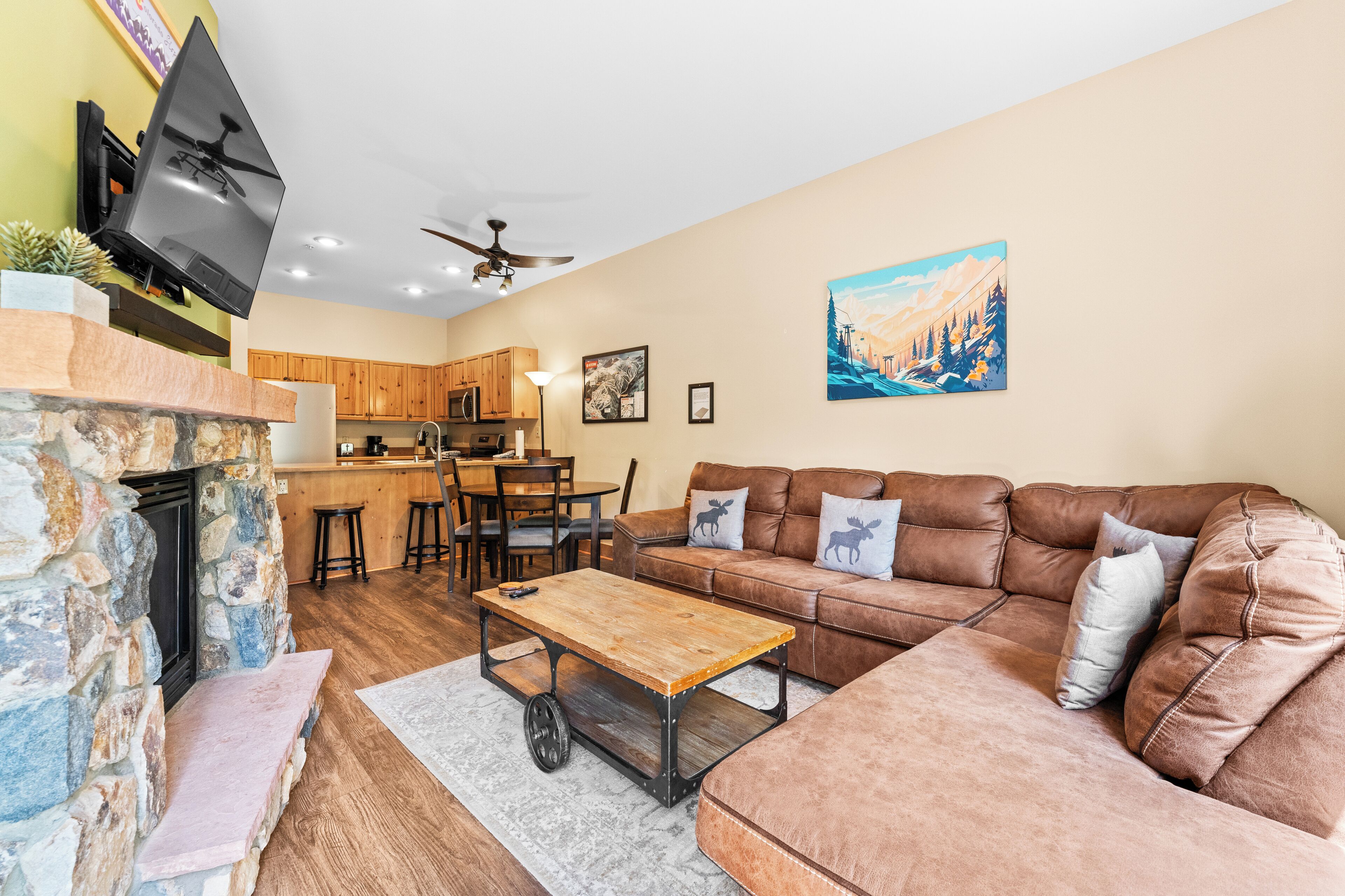 A living room with a stone fireplace, a brown leather sofa, a wooden coffee table, and a kitchen in the background.