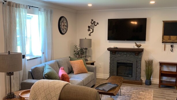 Una sala de estar con sofá, sillón, mesa de centro, chimenea, televisión y un reloj en la pared.