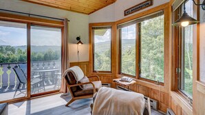 Ein gemütliches Zimmer mit Holzbalkendecke, großen Fenstern mit Blick ins Freie, einem Sessel, einem kleinen Tisch und einem Bett.