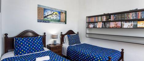 Une chambre avec deux lits, une table de chevet, une lampe, une bibliothèque et un tableau au mur.