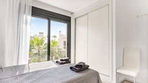 Una camera d'hotel con un letto, una sedia, una finestra con tende e vista su palme e palazzi.