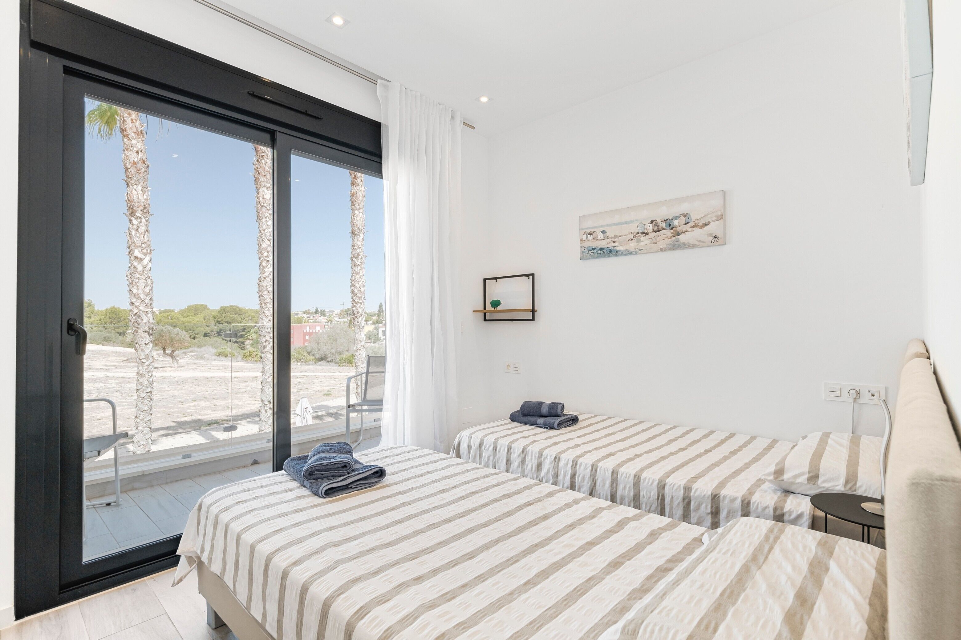 Een kamer met twee bedden, een raam met uitzicht op palmbomen, een schilderij aan de muur en een klein tafeltje.