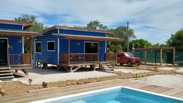 Una piscina con cubierta de madera, un coche rojo aparcado cerca y un edificio azul con porche.