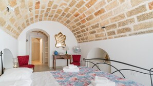 A bedroom with a brick ceiling, a bed with floral bedding, a red chair, a bedside table with a lamp, and a mirror on the wall.