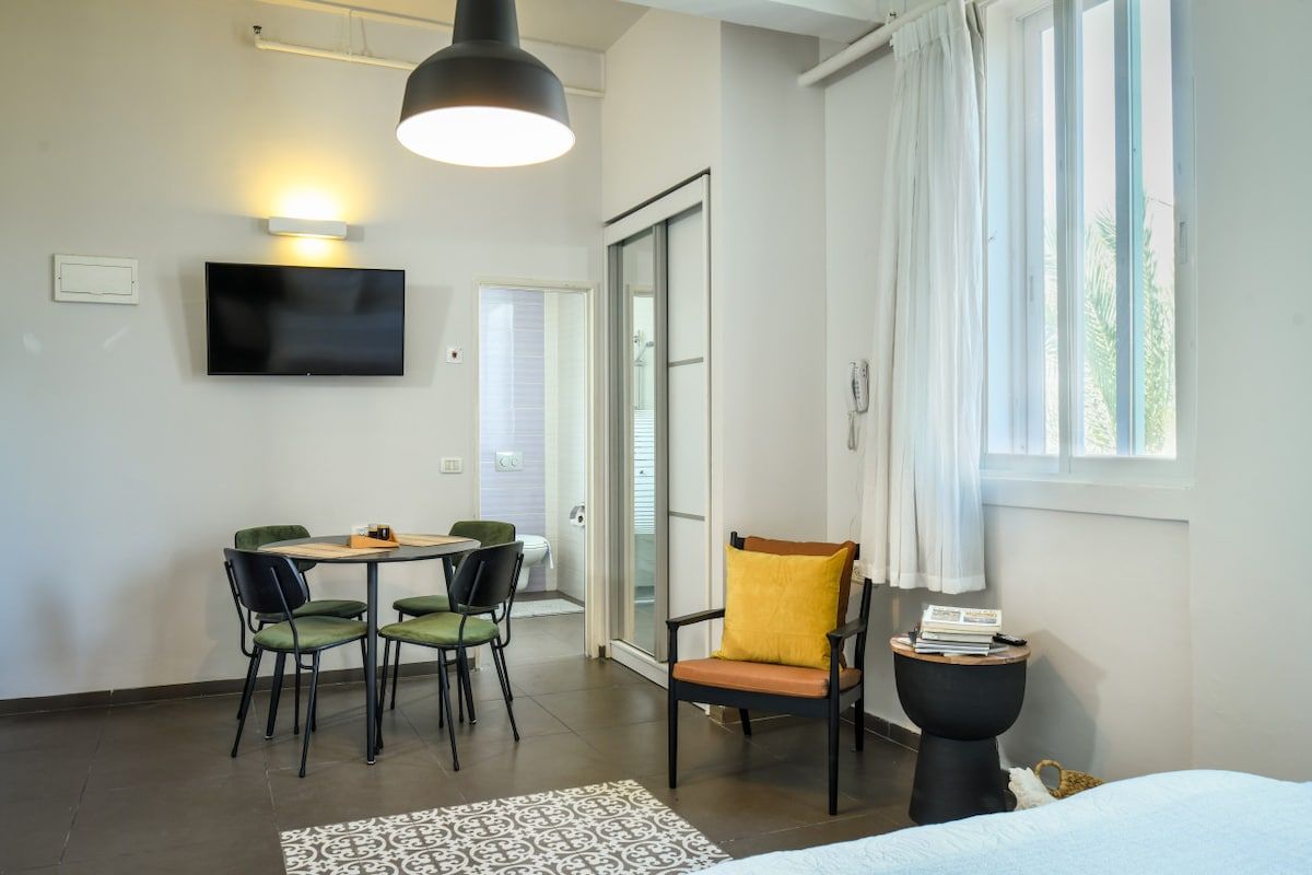 A modern hotel room with a bed, a dining area with chairs, a television, and a large window with curtains.