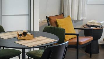 Una habitación con una mesa redonda, dos sillas, una mesita auxiliar con libros y una ventana con cortinas.