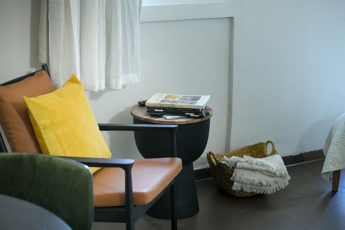Una habitación con una silla, una mesita con libros, una cesta y una ventana con cortinas.