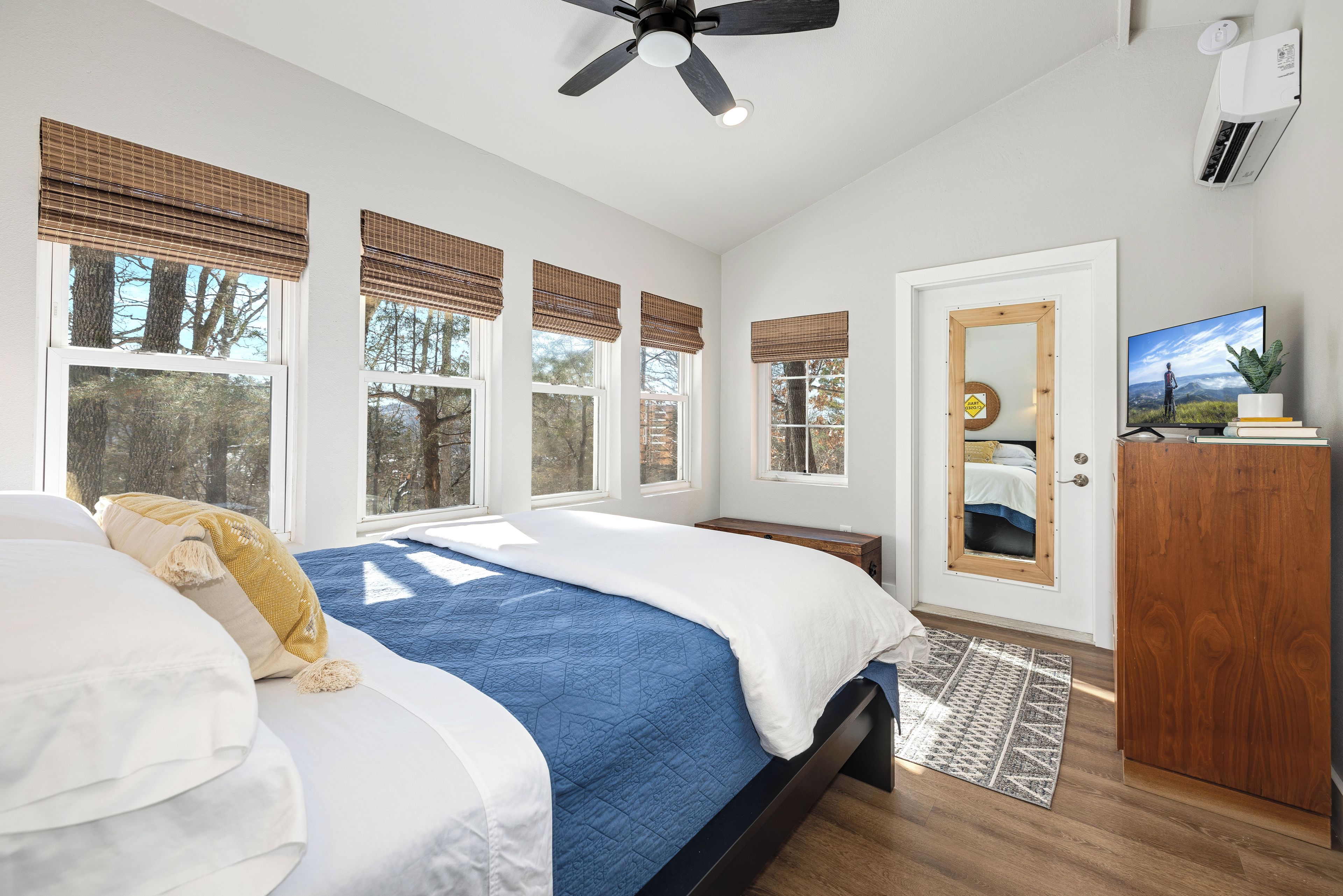 A bedroom with a bed, a ceiling fan, a TV, and a window with a view of trees.