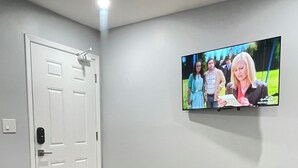 Una sala de estar con un televisor de pantalla plana montado en la pared, una mesa redonda con un control remoto, una pequeña planta en maceta y una alfombra gris.