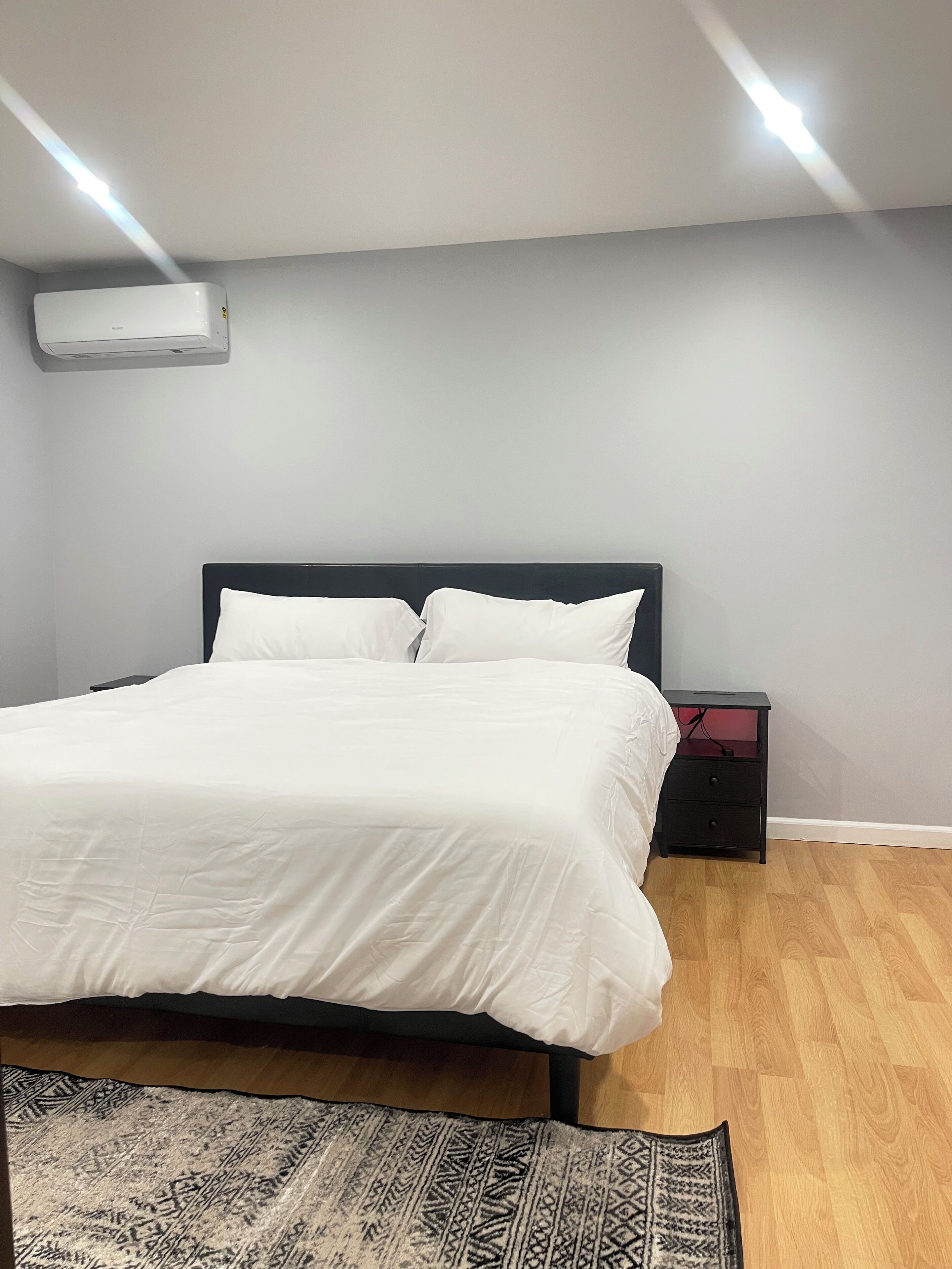 Een keurig opgemaakt bed met witte beddengoed in een kamer met lichtgrijze muren, een houten vloer en een kamerbreed tapijt met dessin.