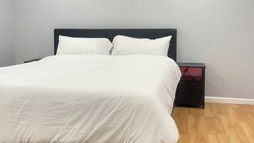 Een keurig opgemaakt bed met witte beddengoed in een kamer met lichtgrijze muren, een houten vloer en een kamerbreed tapijt met dessin.