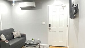 Un espacio habitable compacto con un sofá gris, una pequeña mesa redonda con una planta, una puerta blanca y un televisor montado en la pared.